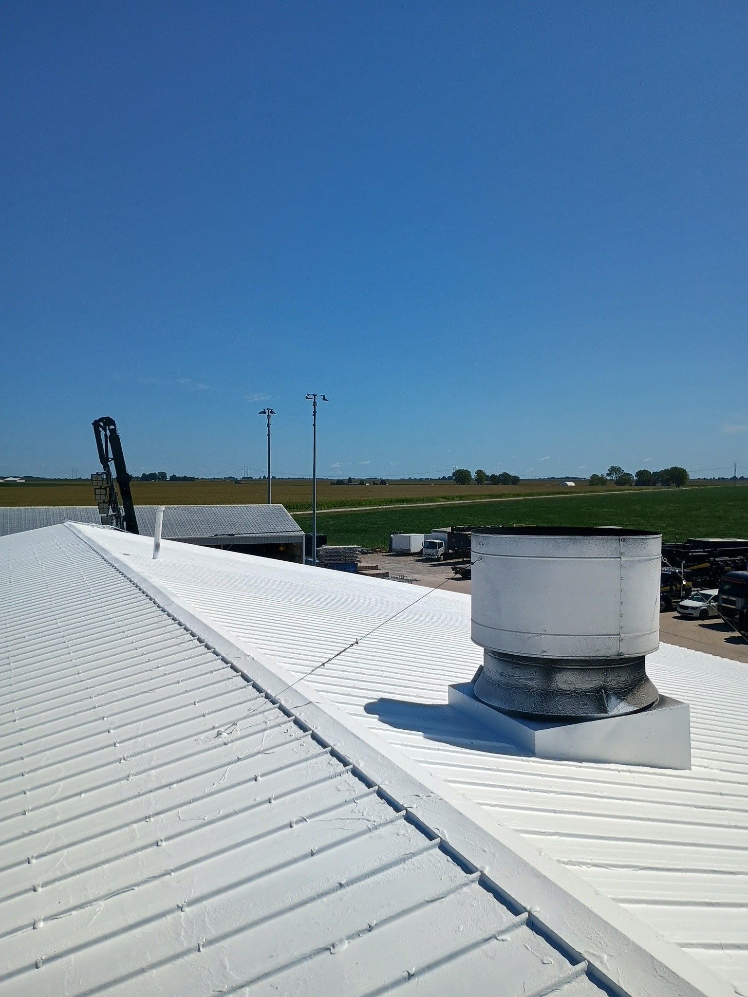 White metal rooftop with a vent under a clear blue sky, viewed over open fields.