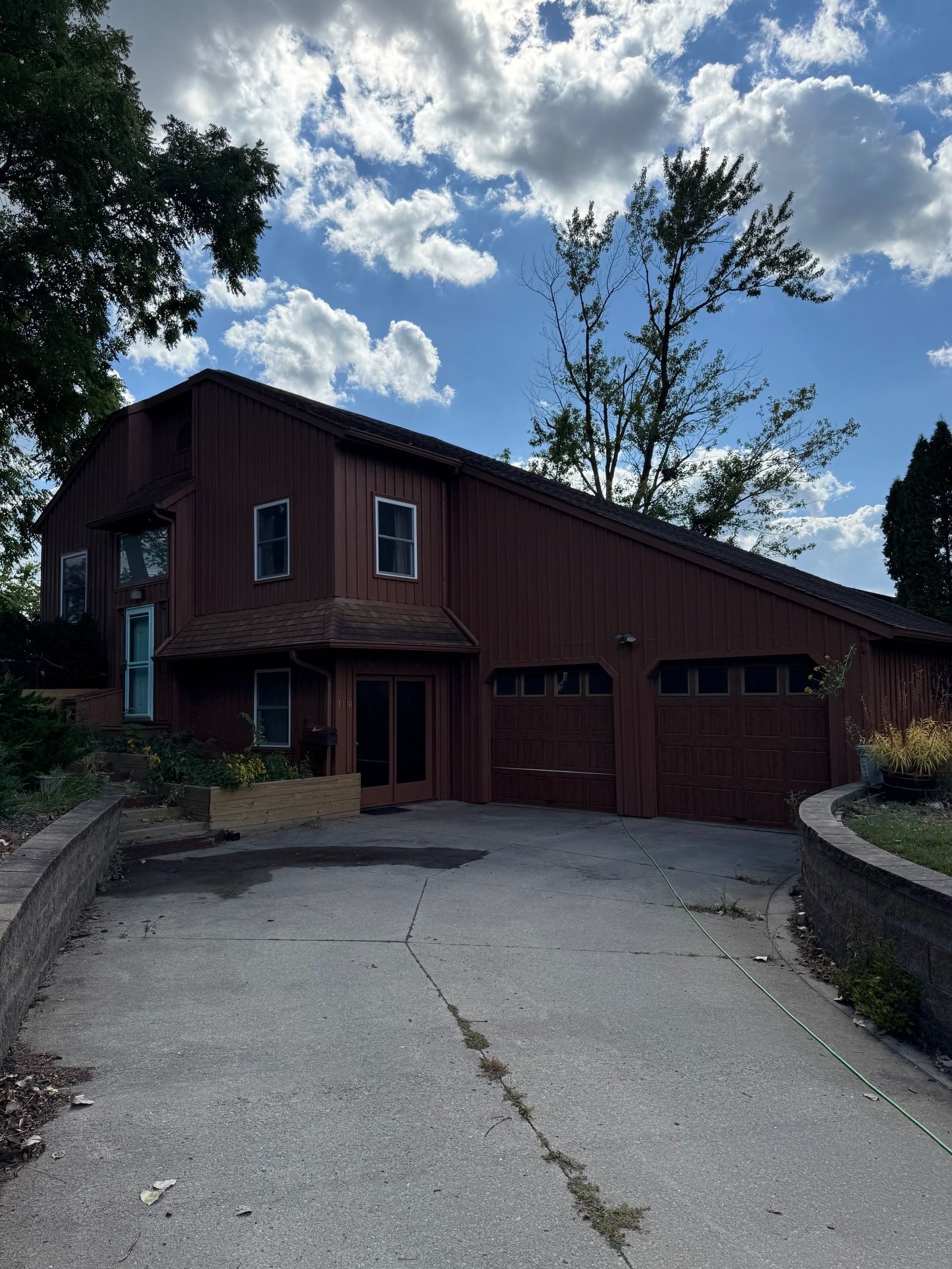 Rustic two-story house with red siding, garage doors, and a curved driveway under a partly cloudy sky