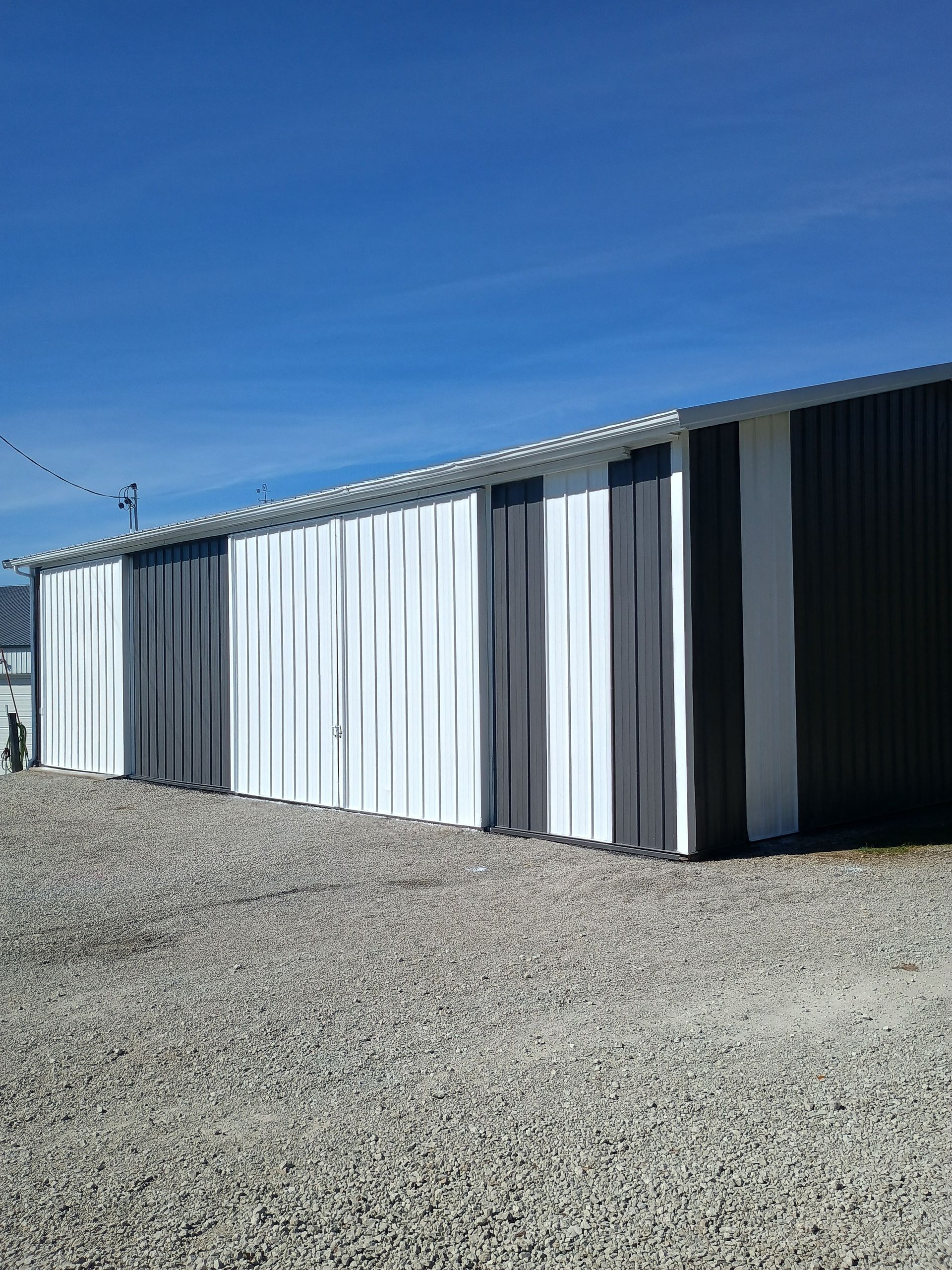 A long, metal-sided building with alternating vertical panels of white and dark gray sits on a gravel lot under a blue sky.