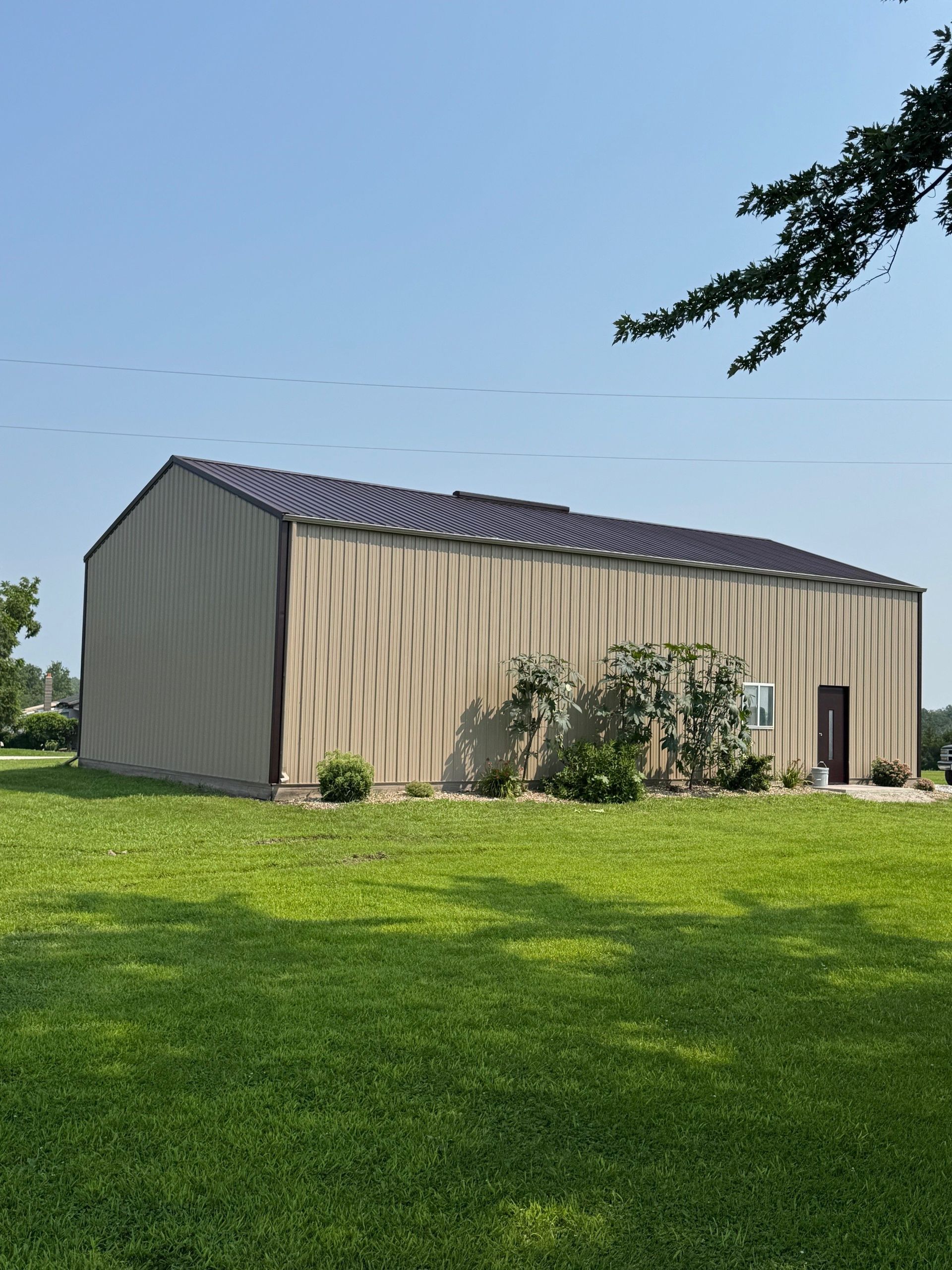 A tan metal building with a dark roof sits on a green, grassy lawn under a clear blue sky.