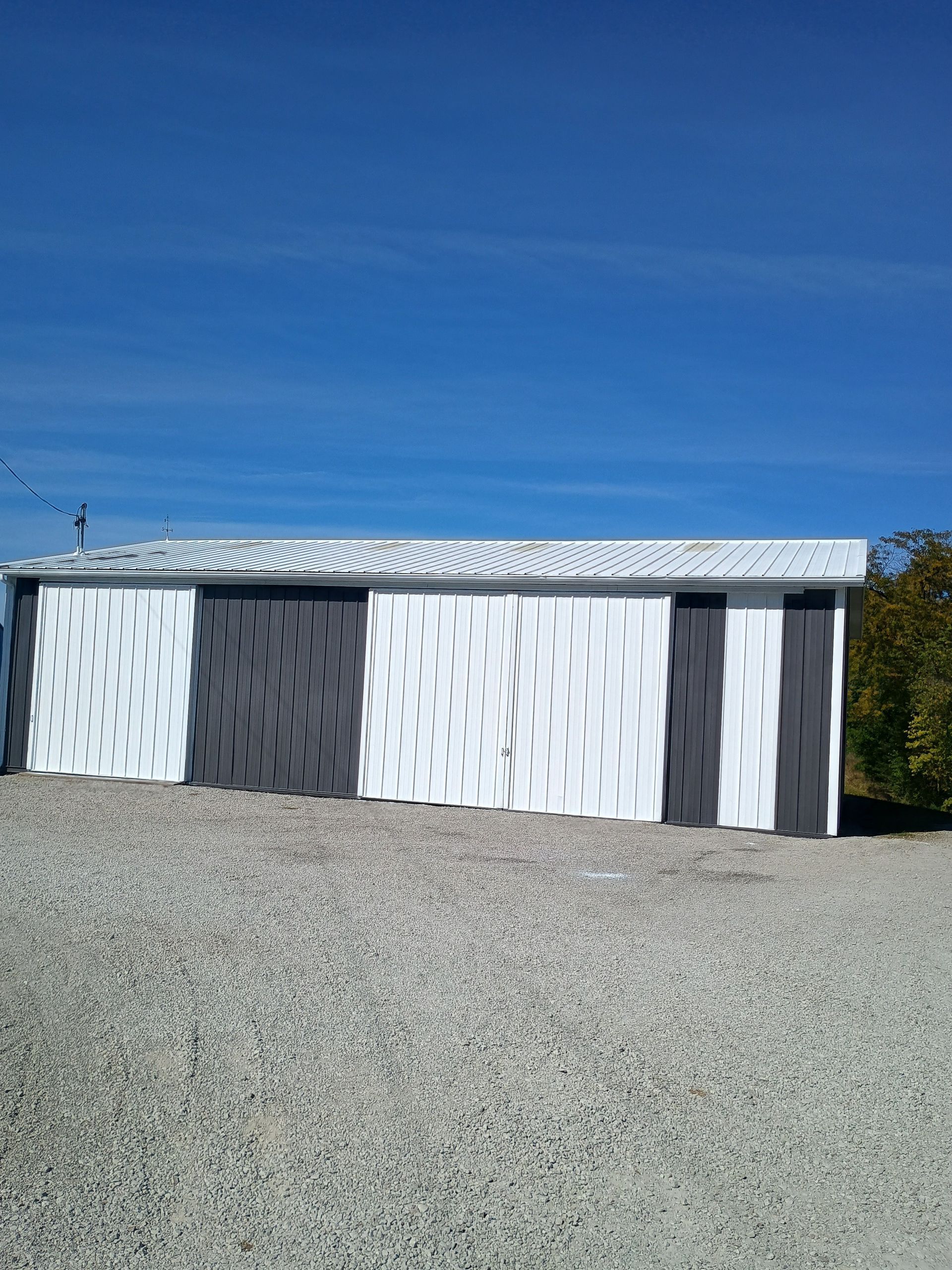 A long storage shed with alternating black and white vertical panels stands on a gravel lot under a clear blue sky.