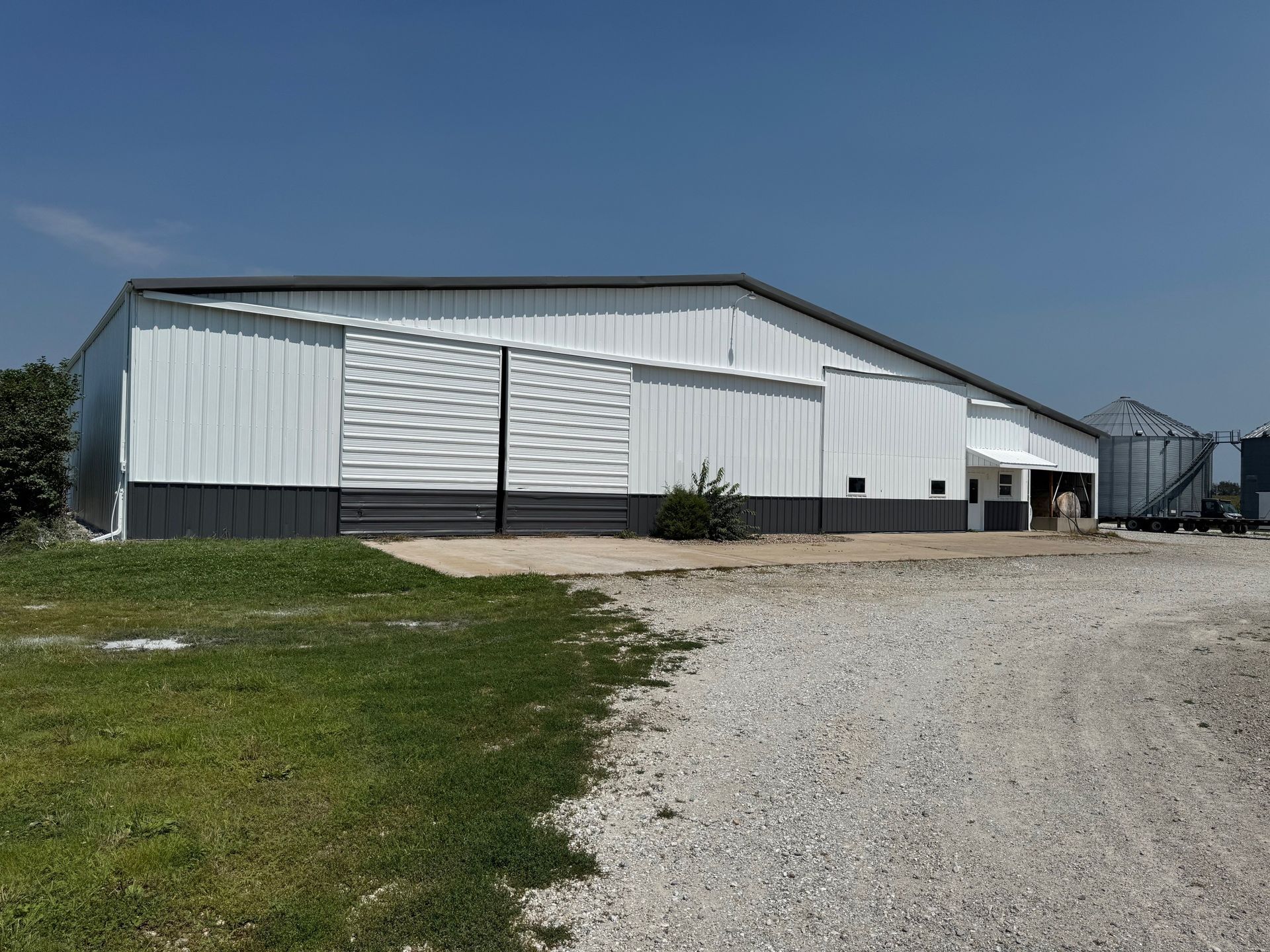 A large white agricultural metal building with a dark gray base, set against a blue sky, next to a gravel path and lawn.