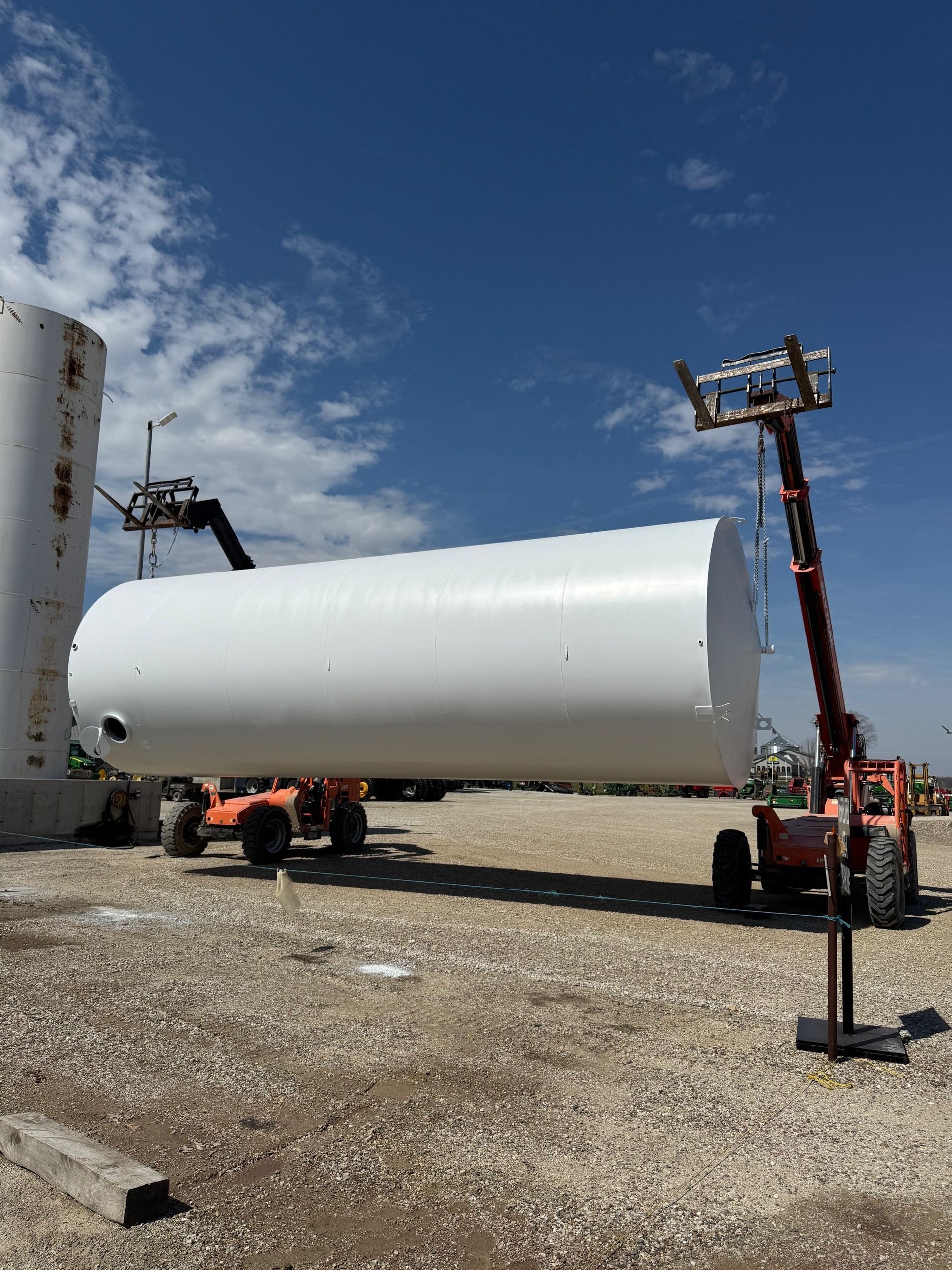 Two orange telehandlers lift a large white cylindrical tank on a gravel construction site under a blue sky.