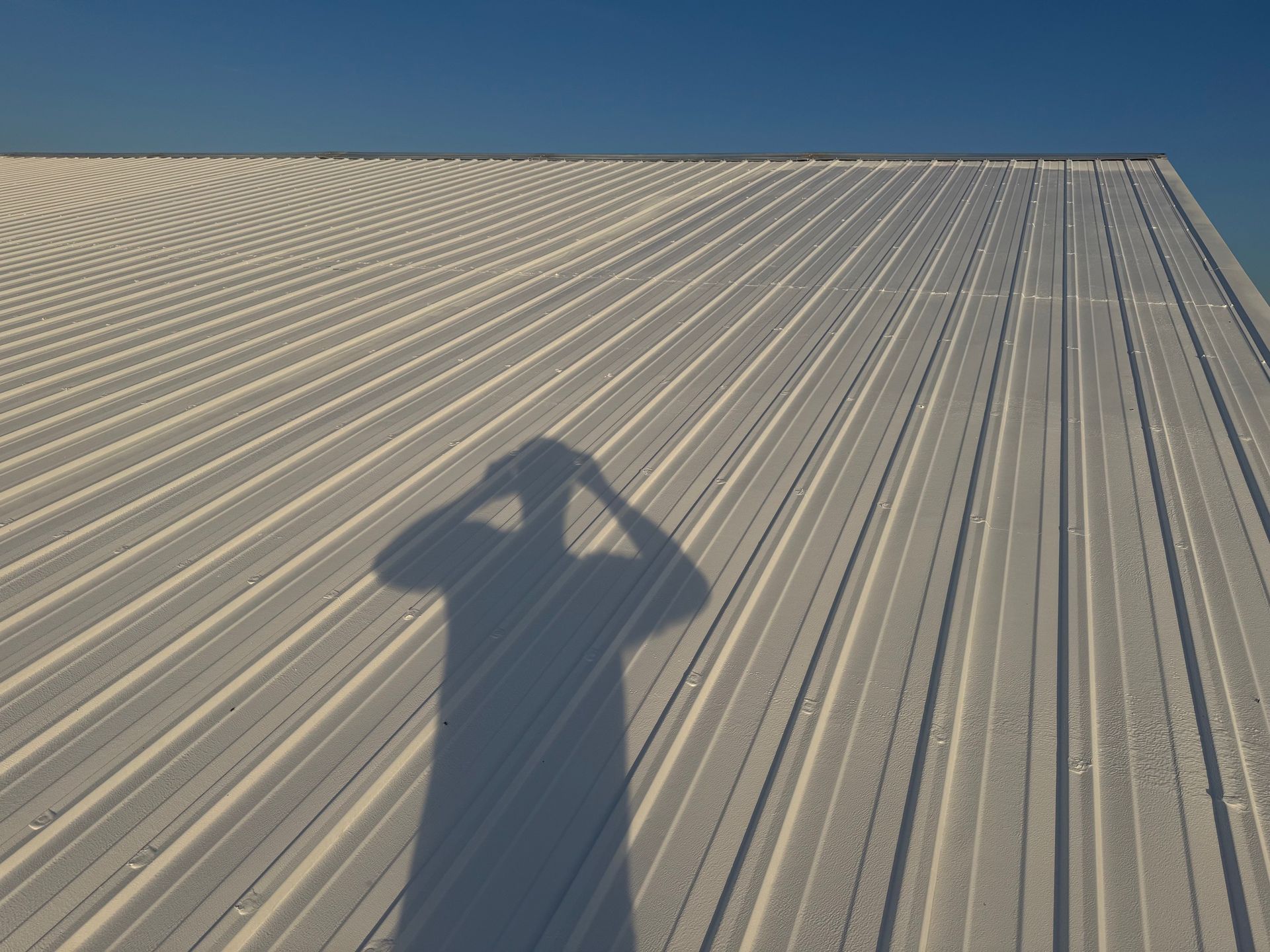 A person's shadow cast onto a bright, white, corrugated metal roof under a clear blue sky.