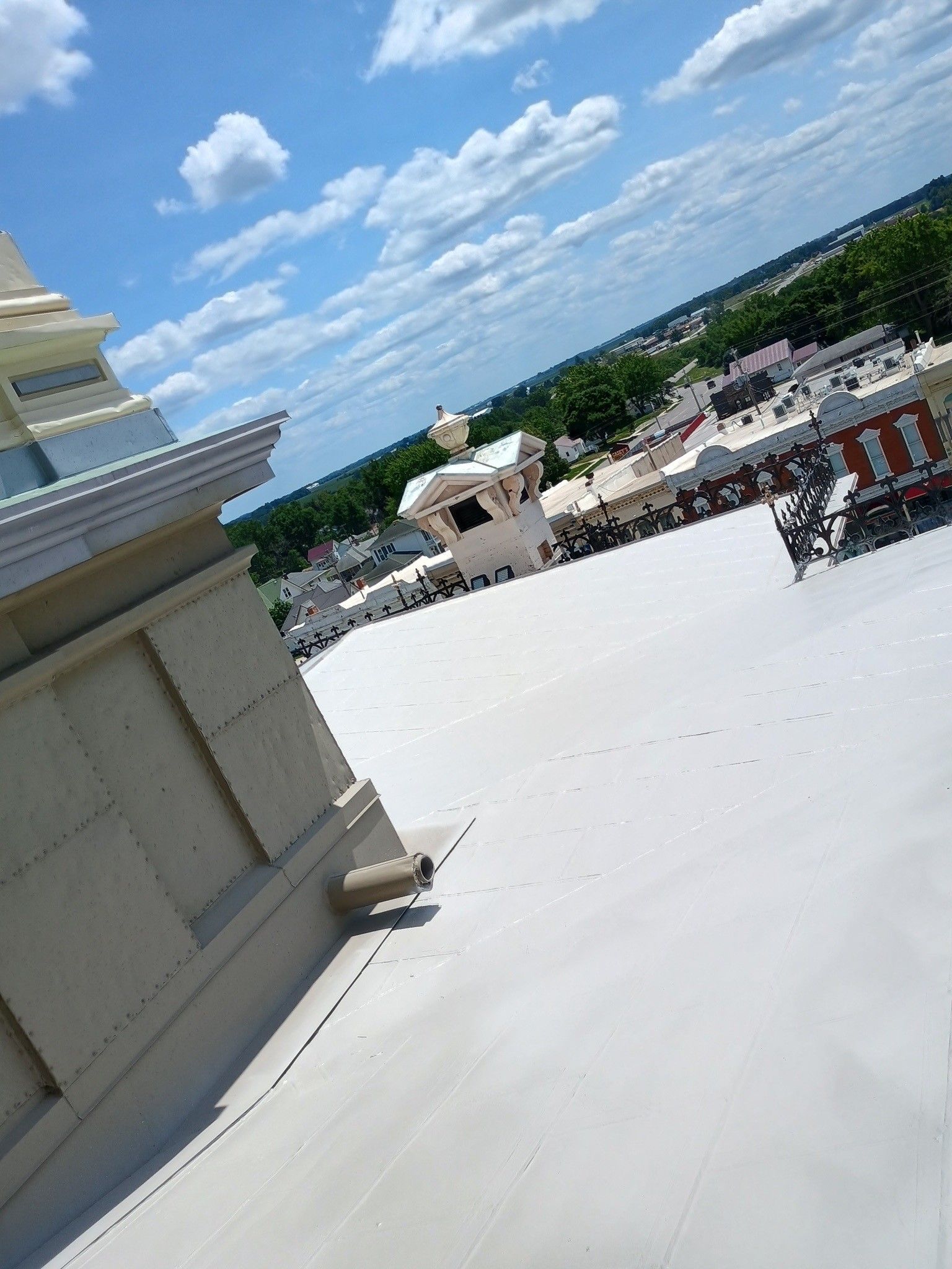 A view from a high rooftop looking over a white flat roof toward a decorative building structure and distant trees.