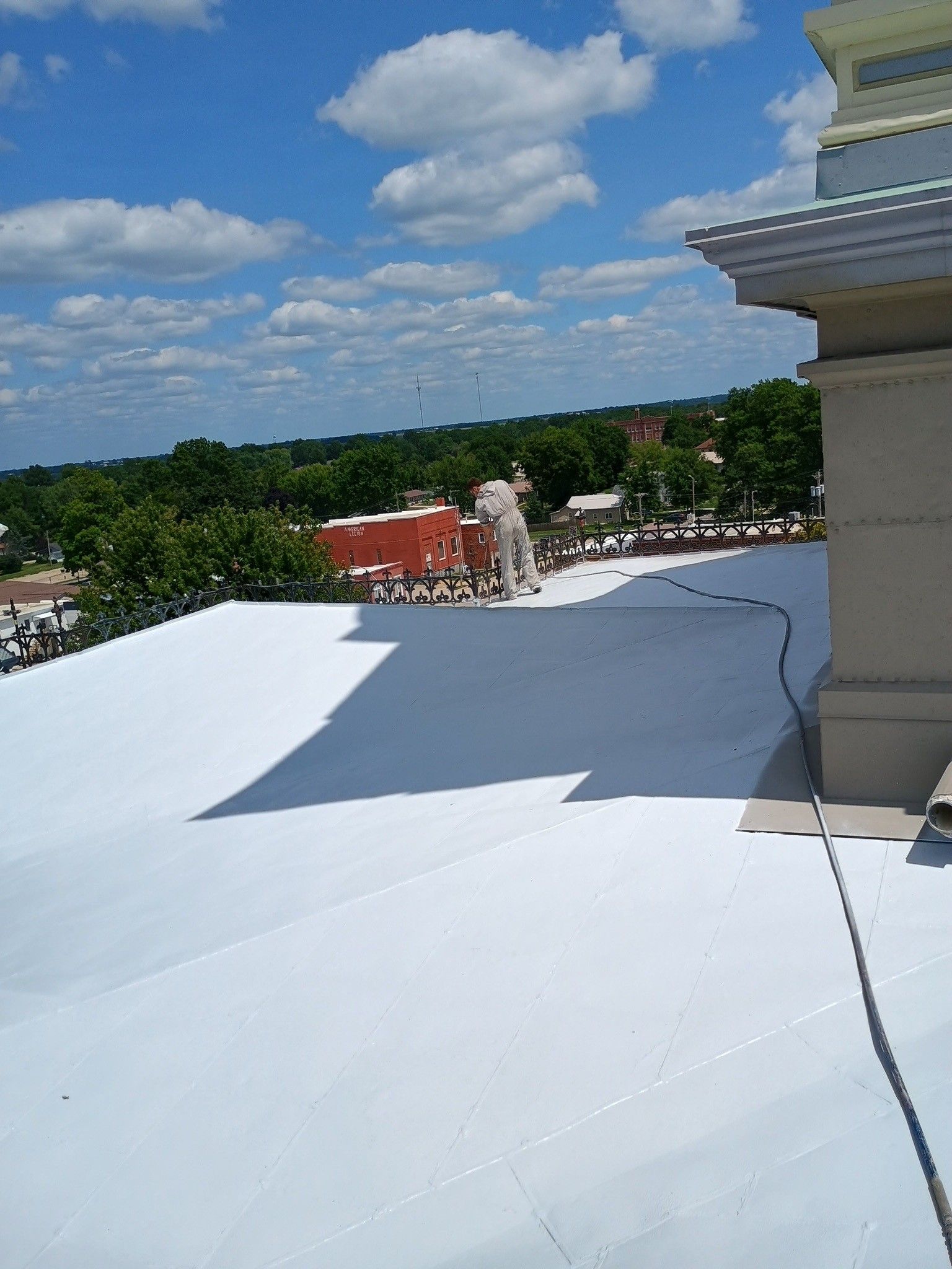 A person in white protective gear spray-paints a large, white flat roof under a blue, cloudy sky.