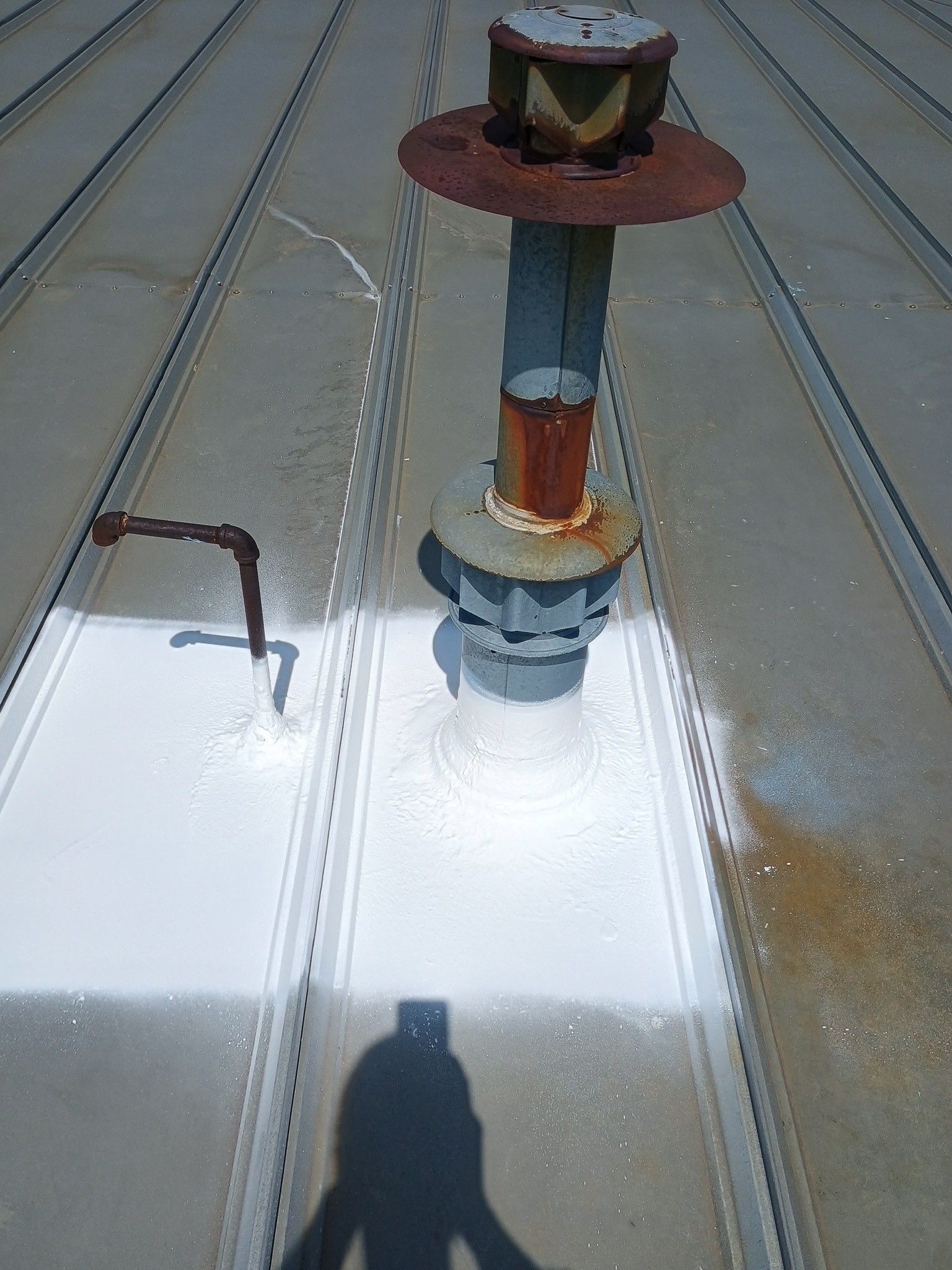 A metal roof with white sealant applied around a pipe vent and a small pipe stub, with a person's shadow at the bottom.