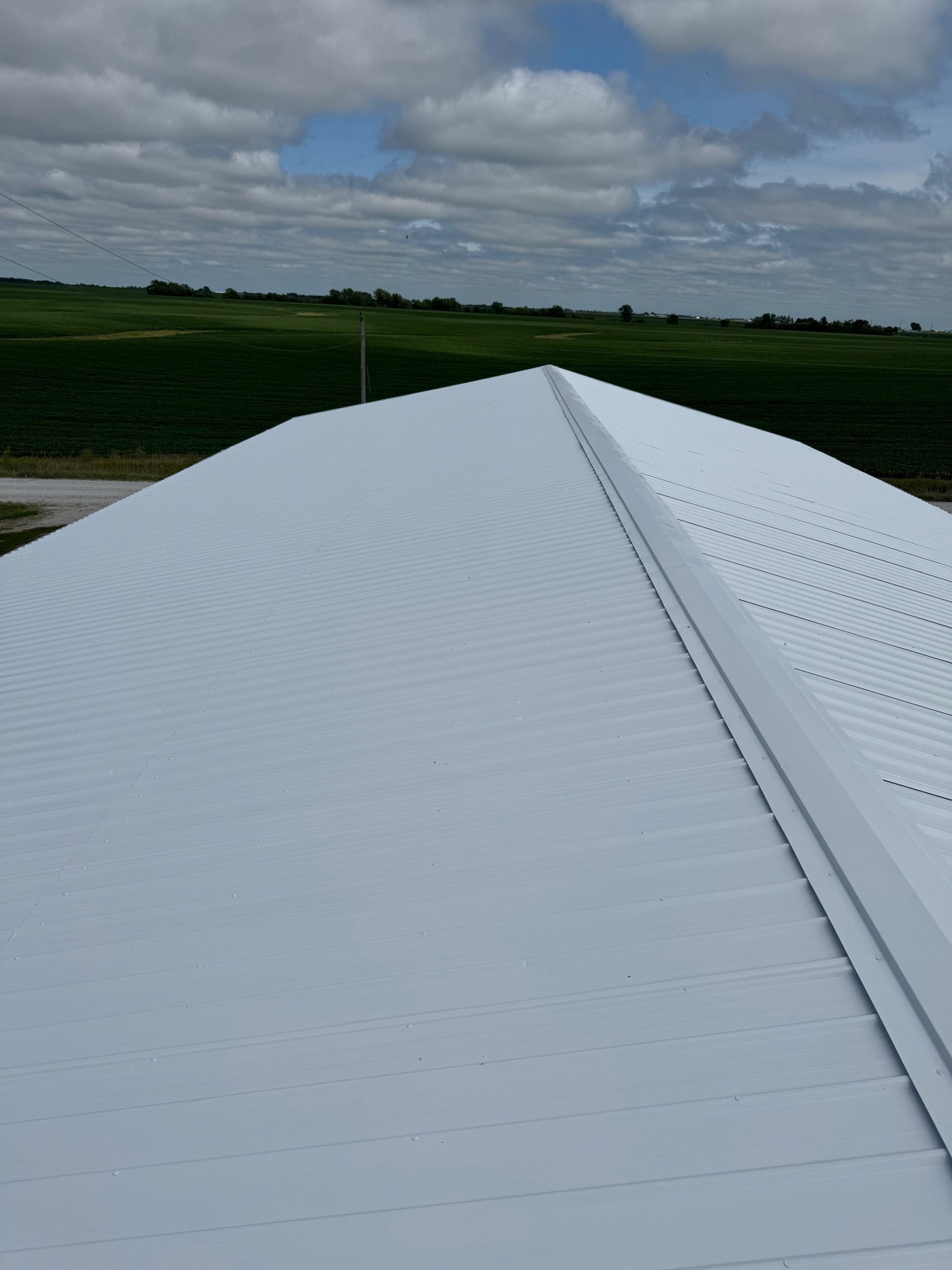 A view looking down the peak of a light gray, corrugated metal roof toward a green field under a cloudy blue sky.