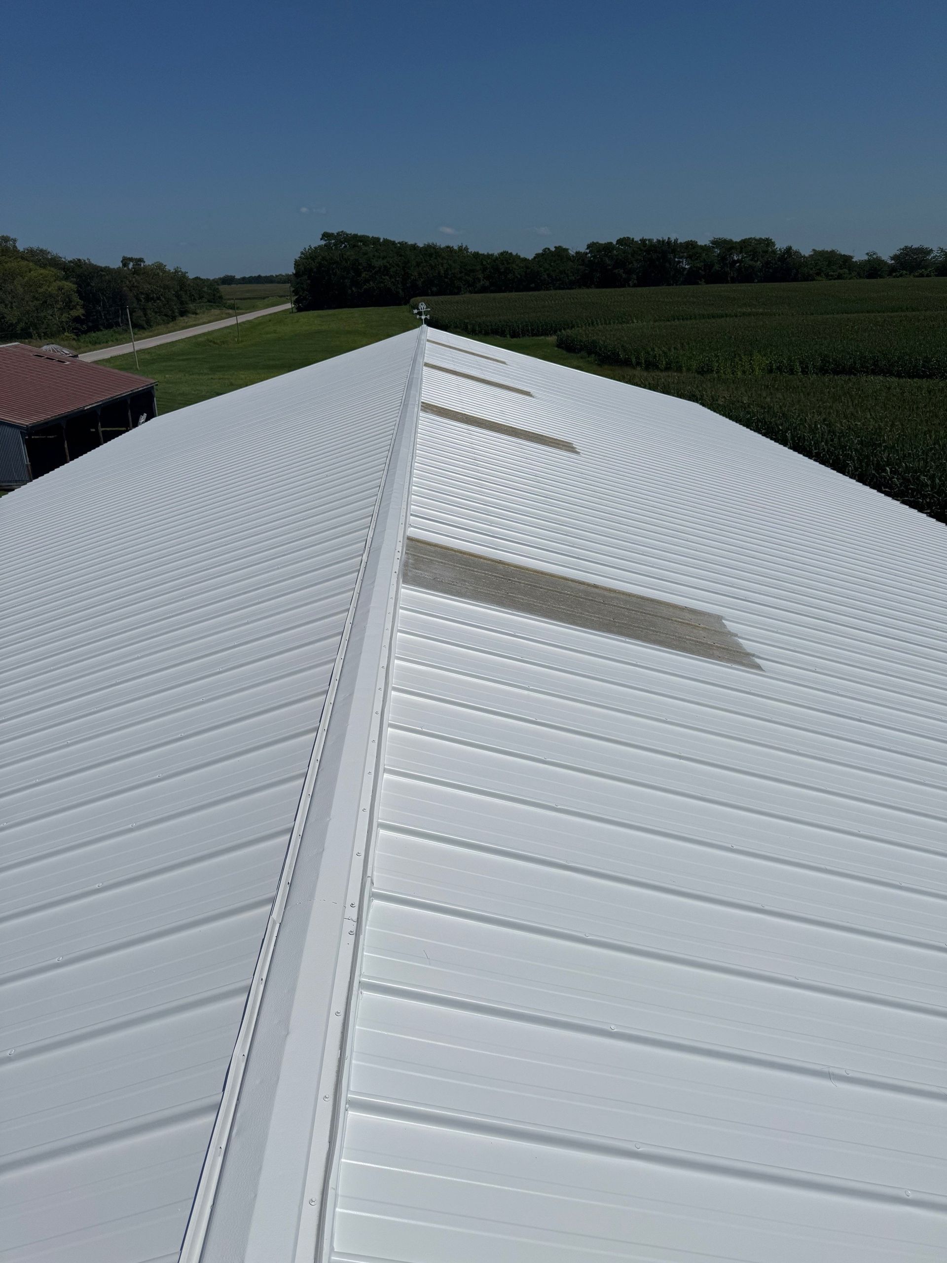 A white, metal-roofed building peak with translucent skylight panels, set against a rural landscape under a clear sky.