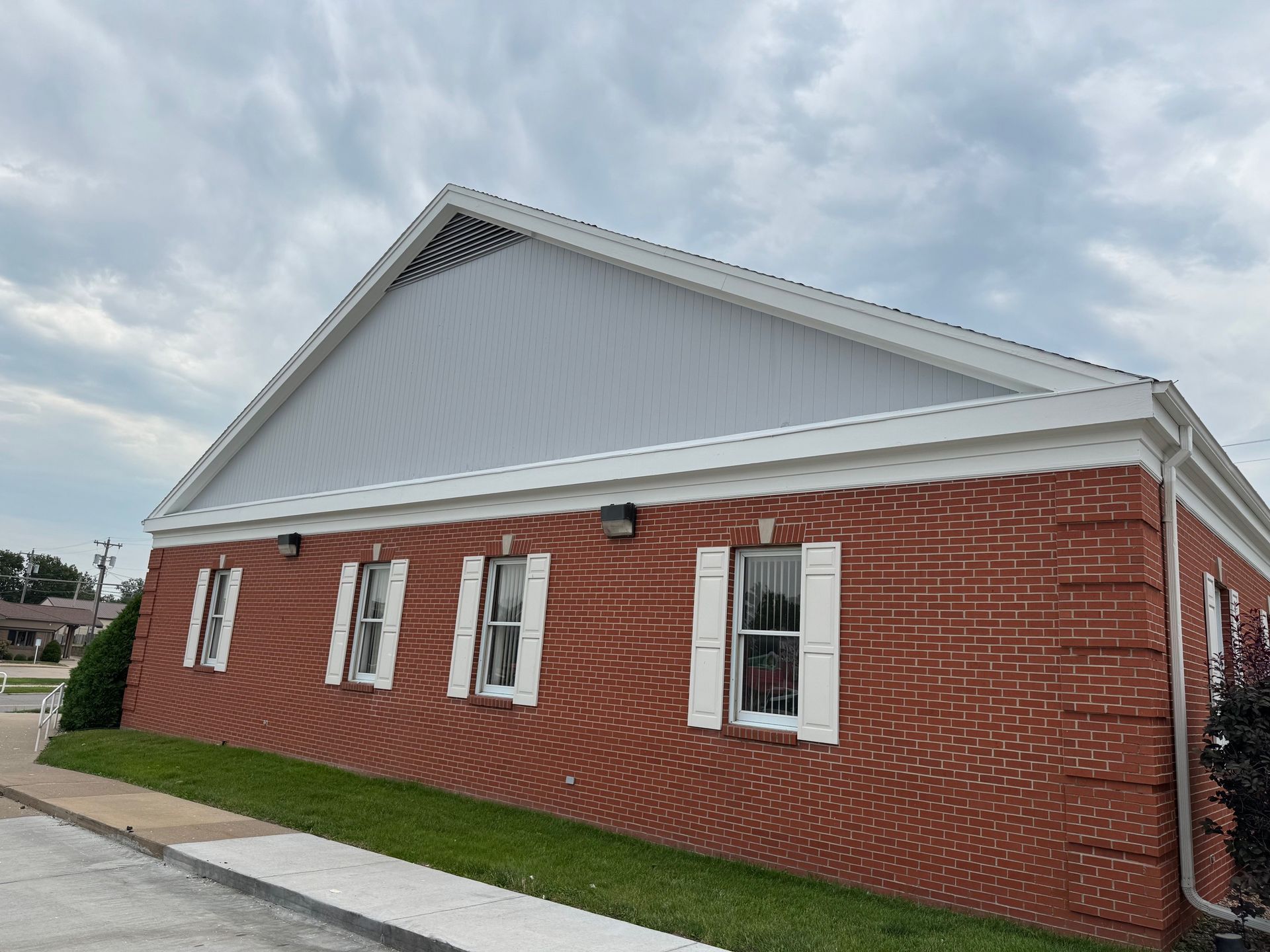 A side view of a one-story brick building with white window shutters and a light gray gable roof under a cloudy sky.