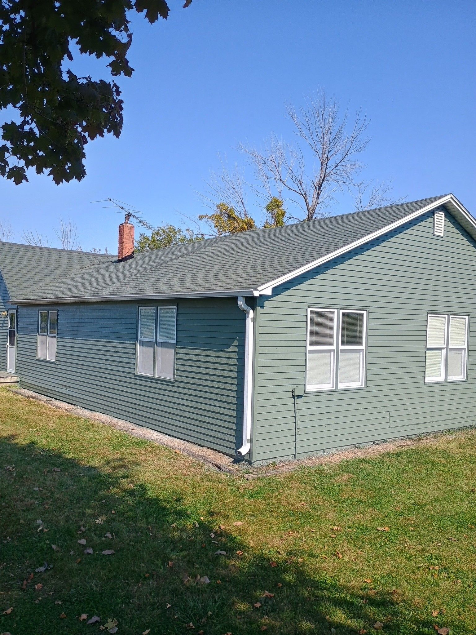 A side view of a house with green siding, a grey shingled roof, a brick chimney, and white-framed windows on a lawn.
