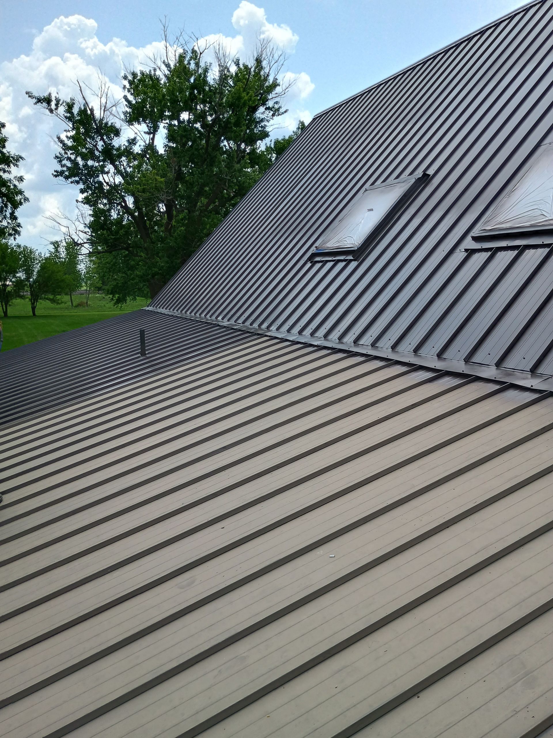 A tan-colored metal roof with vertical seams sits under a clear sky, transitioning to a dark roof section with skylights.