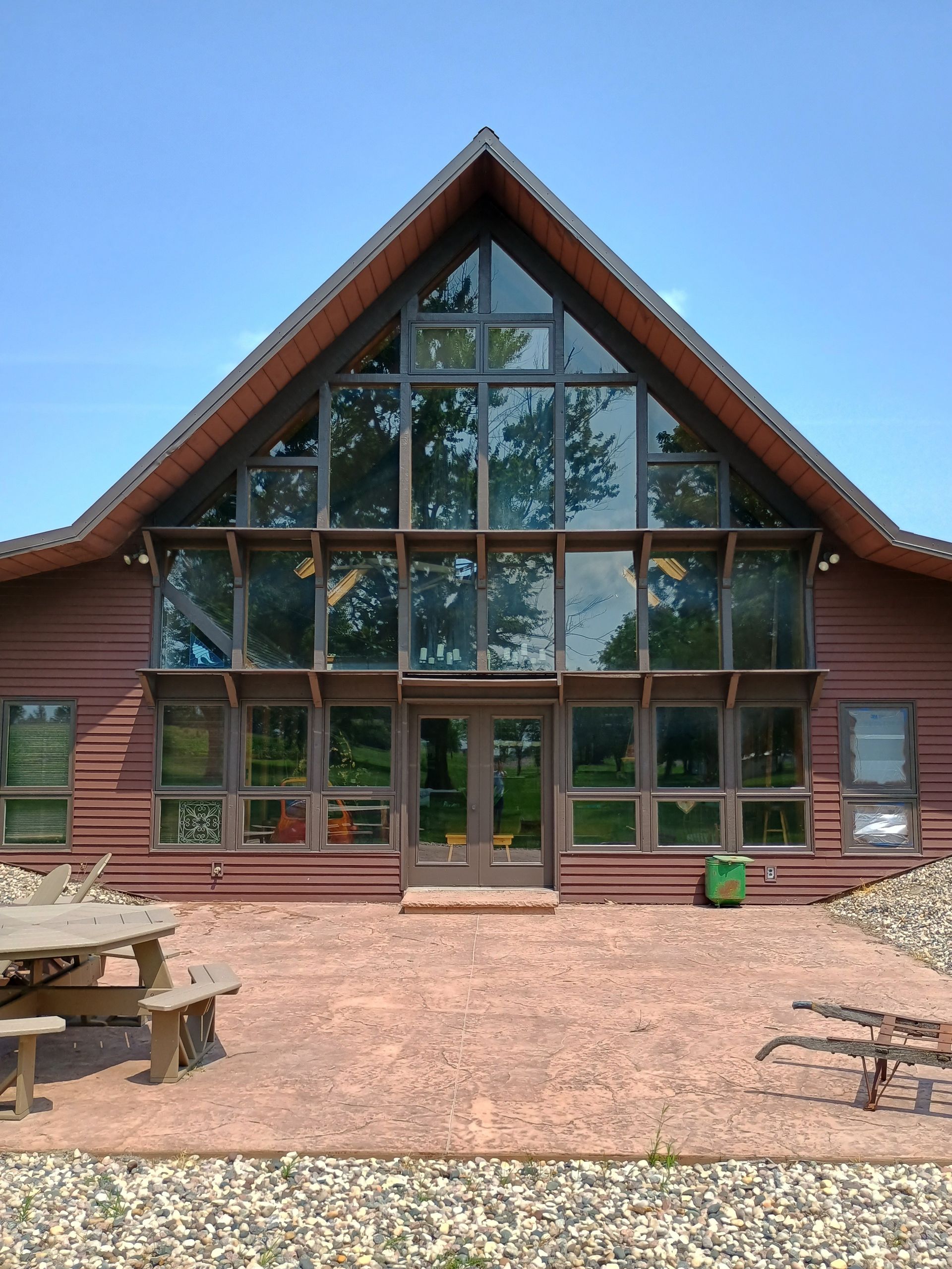 A wooden lodge with a large glass A-frame facade, viewed from a gravel courtyard under a blue sky.