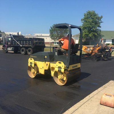 A man is driving a roller on a road next to a dump truck.