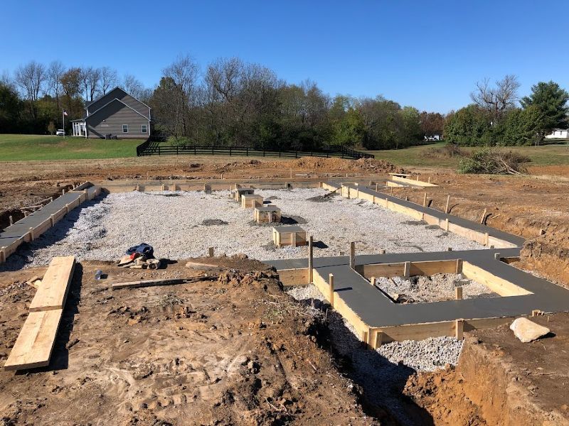 A concrete foundation is being built in a dirt field with a house in the background.