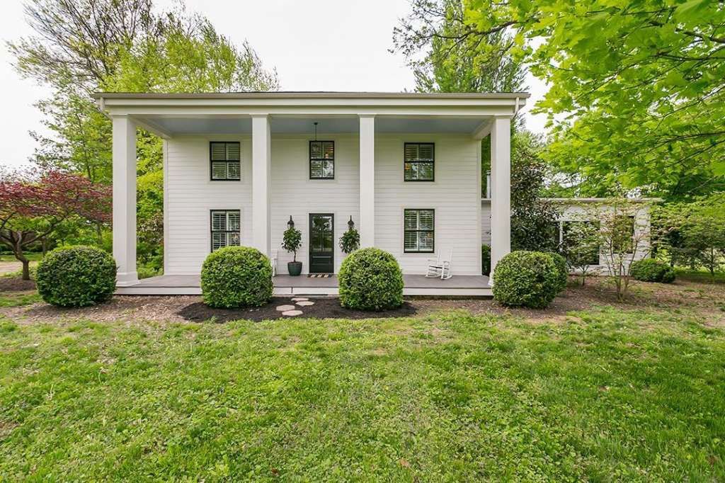 A white house with a porch and bushes in front of it.
