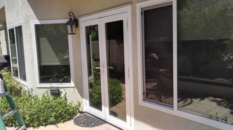 Patio entrance with white double glass doors and windows beside a stucco exterior wall, with plants nearby