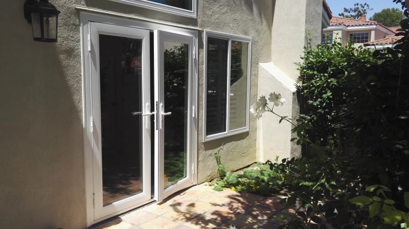 White French doors and a window opening to a sunlit patio beside green shrubs and a beige house wall