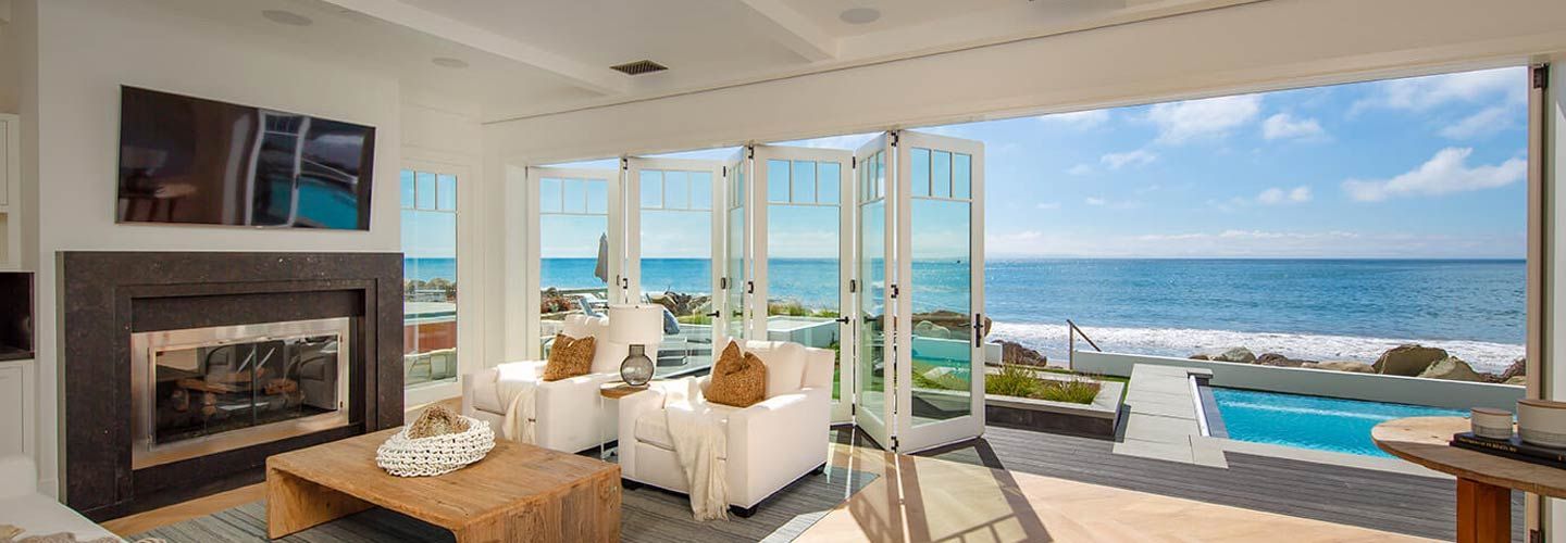 Bright ocean-view living room with fireplace, white seating, and open glass doors to a patio and pool.