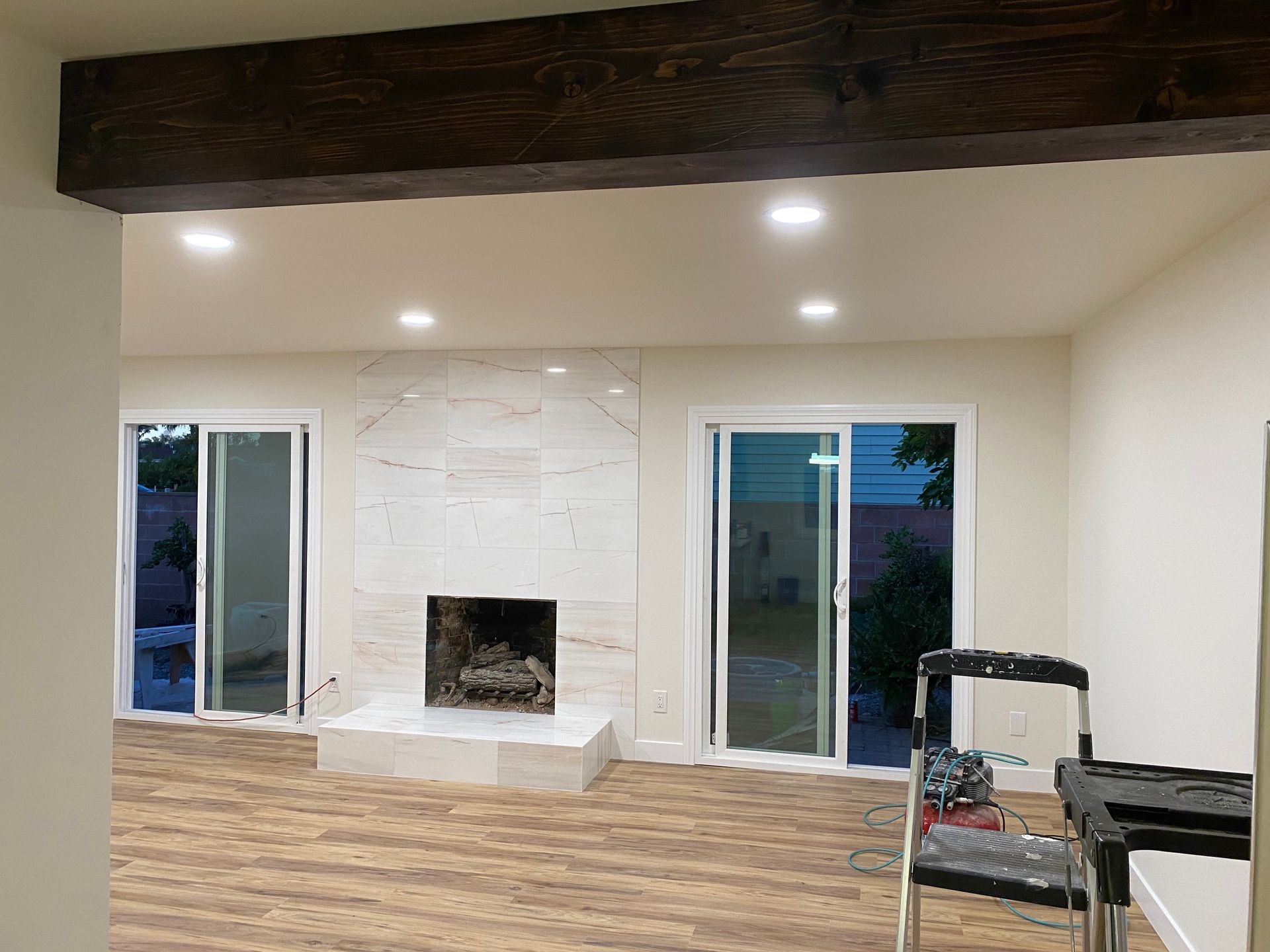 Empty modern living room with white walls, wood floor, exposed beam, recessed lights, fireplace, and sliding glass doors