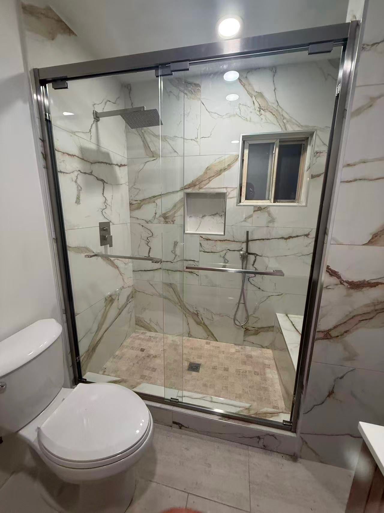 Modern bathroom with a glass walk-in shower, marble-look tile walls, and a toilet in the foreground