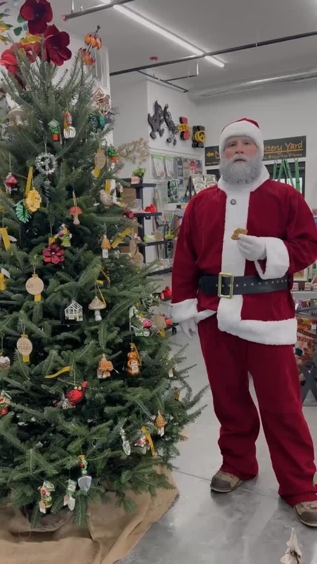 Santa Claus stands by a decorated Christmas tree in a store, holding a cookie.