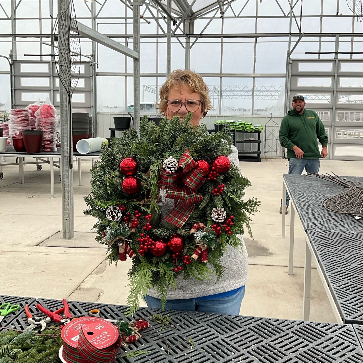 Woman holds a Christmas wreath with red ornaments, indoors at a greenhouse. Another person works in background.