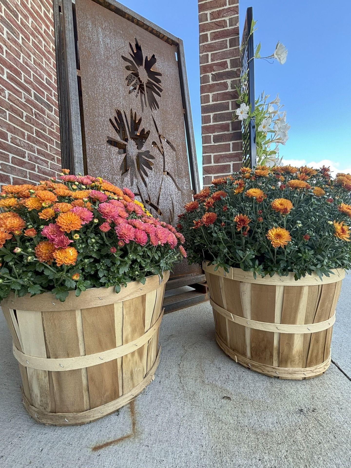 Two large wooden baskets filled with colorful mums against a brick building with a metal floral art piece.