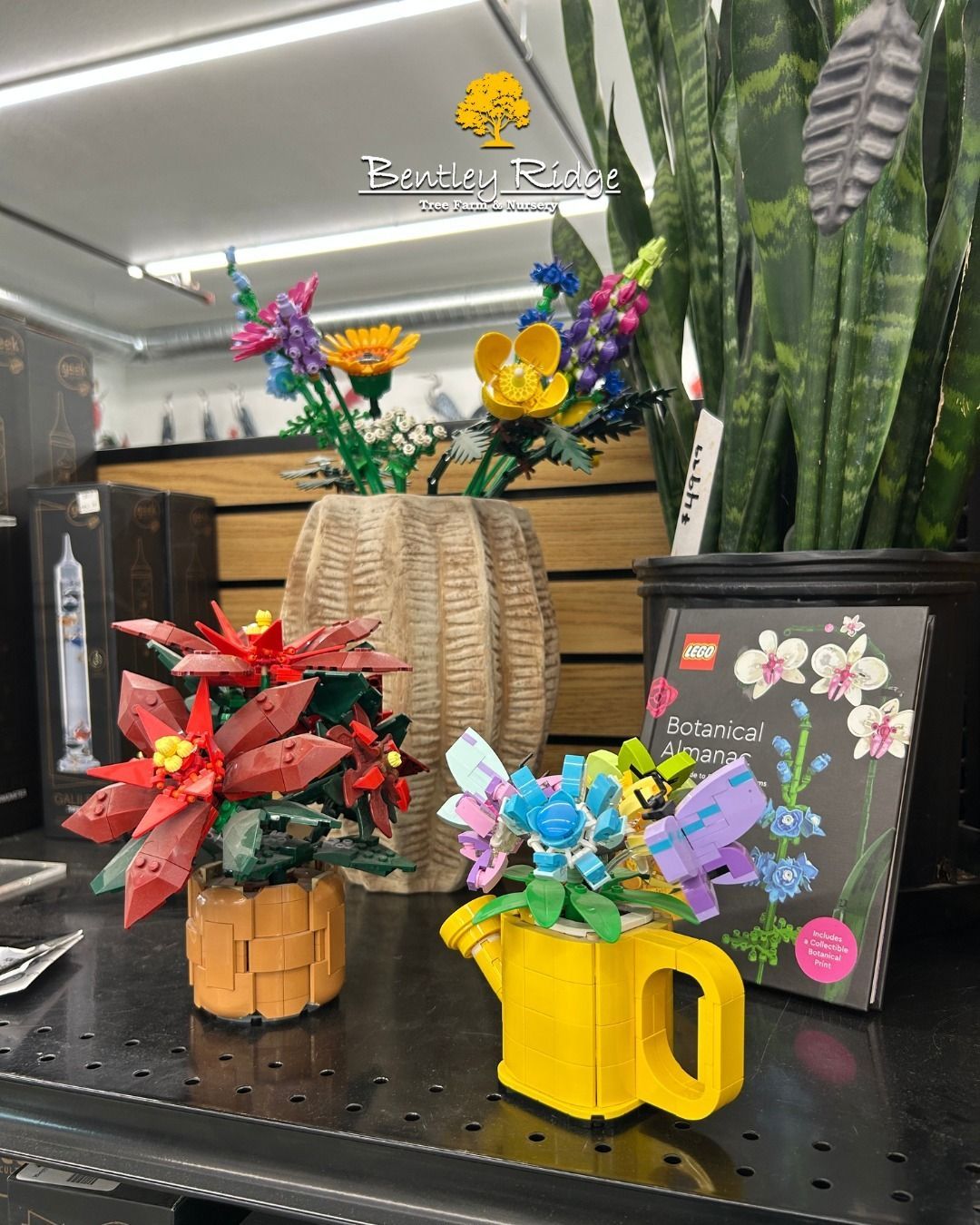 Lego flower sets on display: red, yellow, blue flowers in vases and a watering can, near a book.
