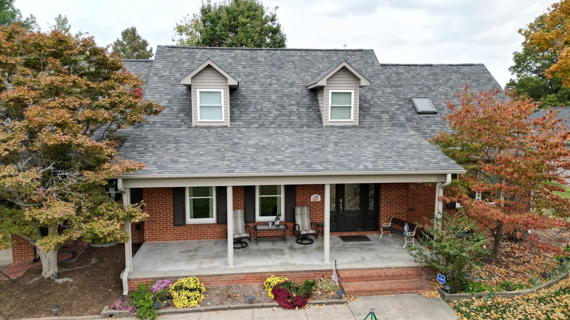 A brick house with a porch, dormers, and colorful autumn trees in front.