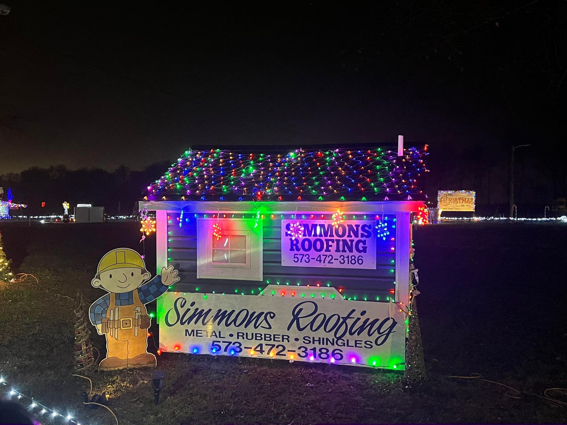 Christmas lights decorate a Simmons Roofing display, including Bob the Builder and a sign with the business's phone number.