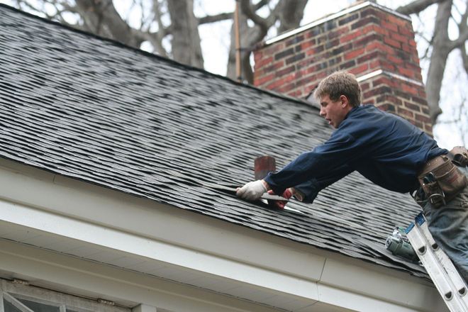 Roofer on a ladder hammering shingles on a dark gray asphalt roof near a brick chimney and white trim.