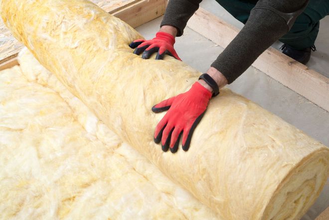 Person in red gloves rolls out insulation between wooden beams.