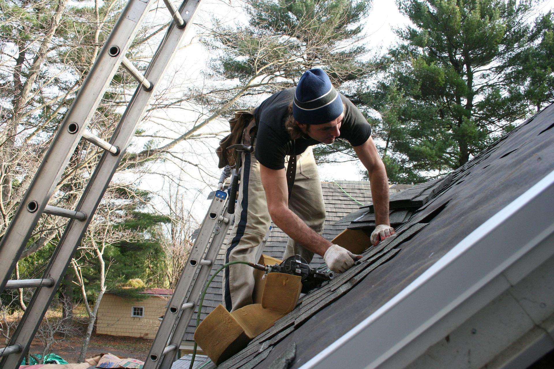 Roofer on a sloped roof replacing shingles near a ladder, wearing work clothes and a beanie.