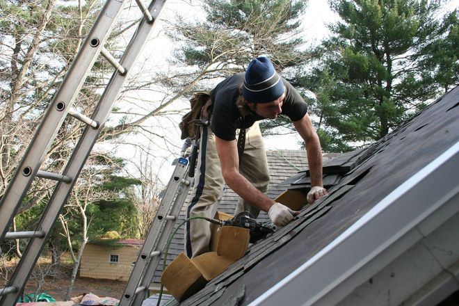 Roofer on a sloped roof replacing shingles near a ladder, wearing work clothes and a beanie.