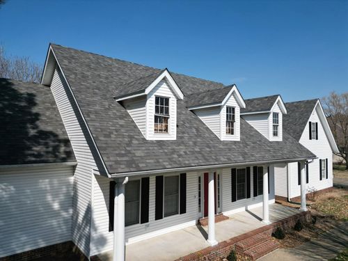 White house with dark gray roof, three dormers, black shutters, and porch.