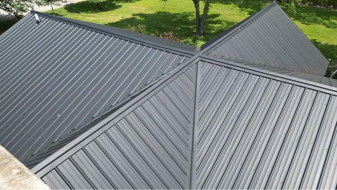 Two roofers in camouflage uniforms installing red metal roofing tiles on a residential roof, blue sky backdrop.