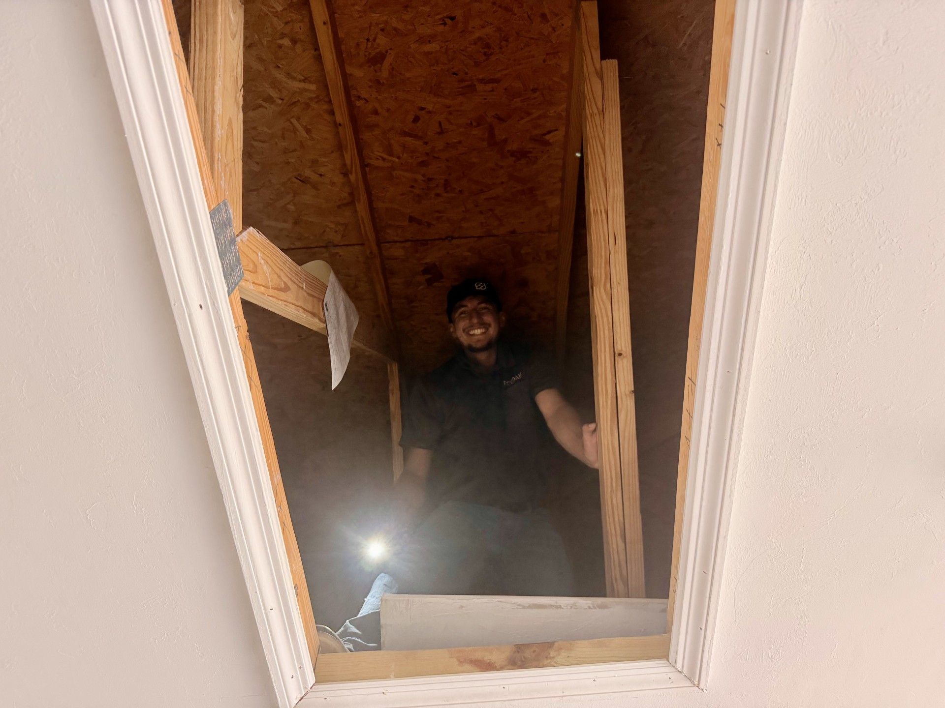 Person standing in an attic opening with a flashlight, framed by exposed wooden rafters.
