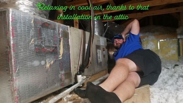 Person lying in a cramped attic beside insulated HVAC ducts and pipes, working on overhead equipment
