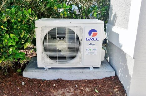 Outdoor Gree air conditioner unit beside a house wall on a concrete pad, with green shrubs nearby