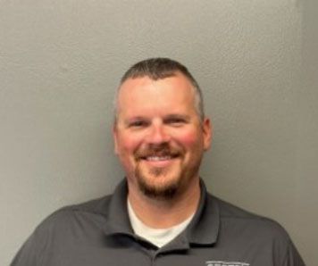 A man with a beard is wearing a gray shirt and smiling for the camera.