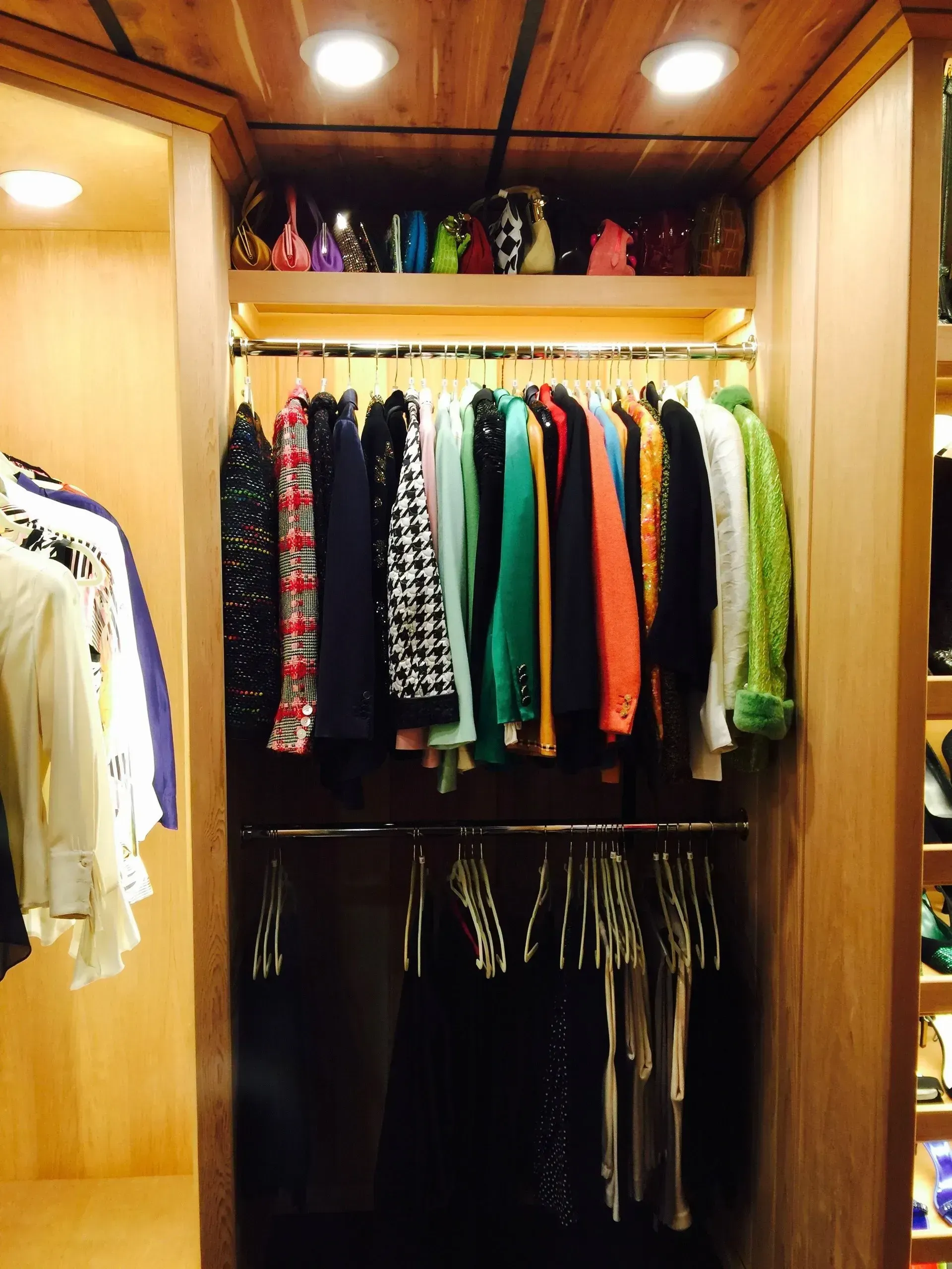 Closet with clothes on hangers and shelves with handbags. Wooden interior with overhead lighting.