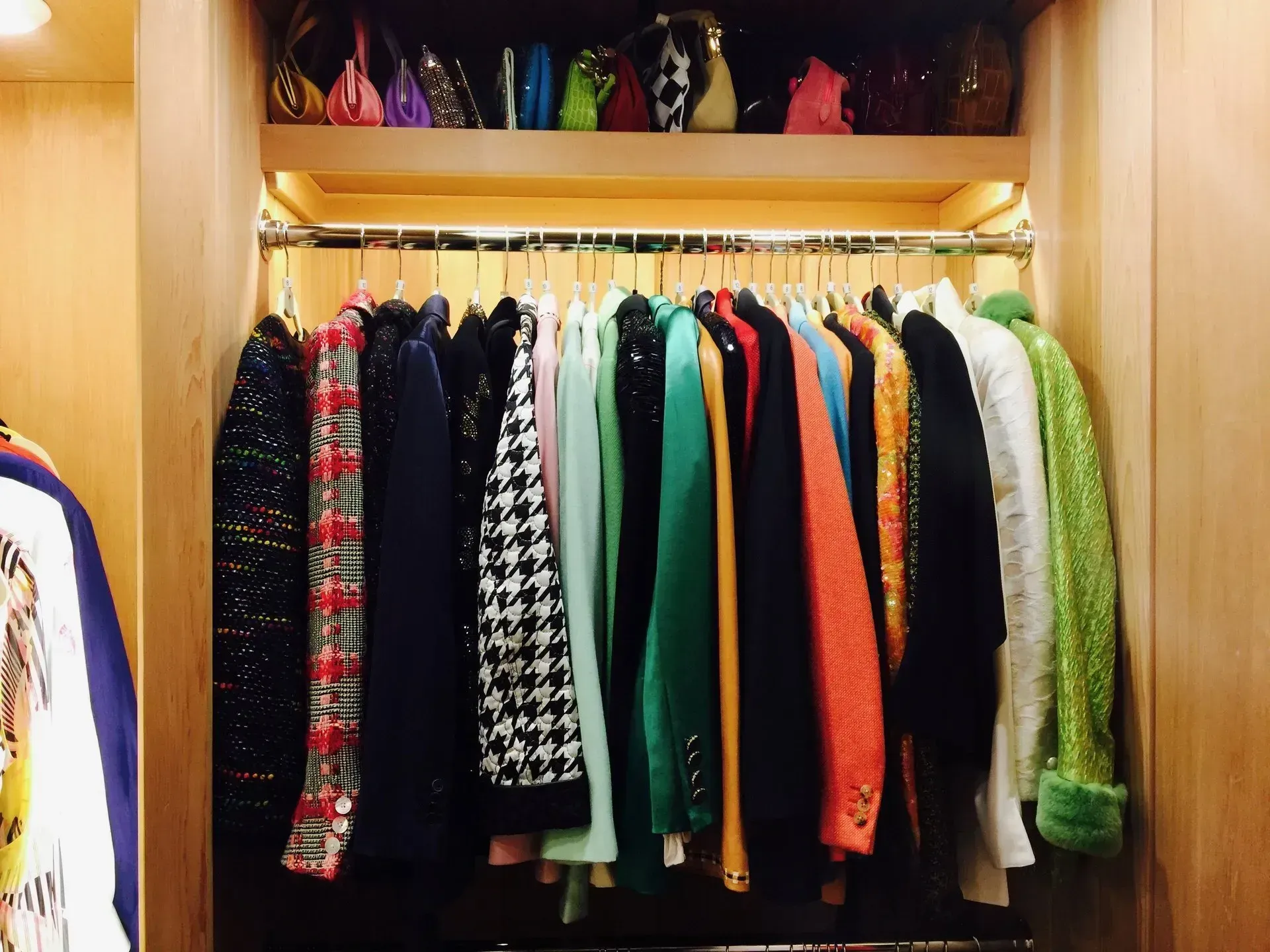 Closet with a clothes rack holding various jackets and coats, and a shelf above with several handbags.