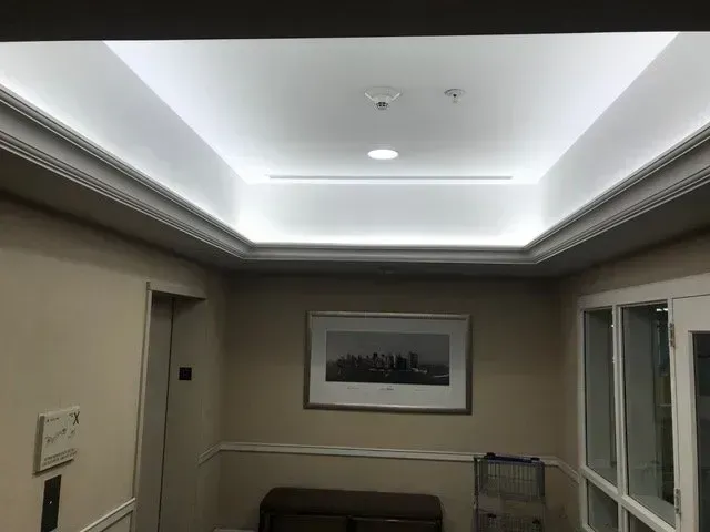 Hallway with recessed, backlit ceiling and artwork. Beige walls, white trim, bench, and double doors.