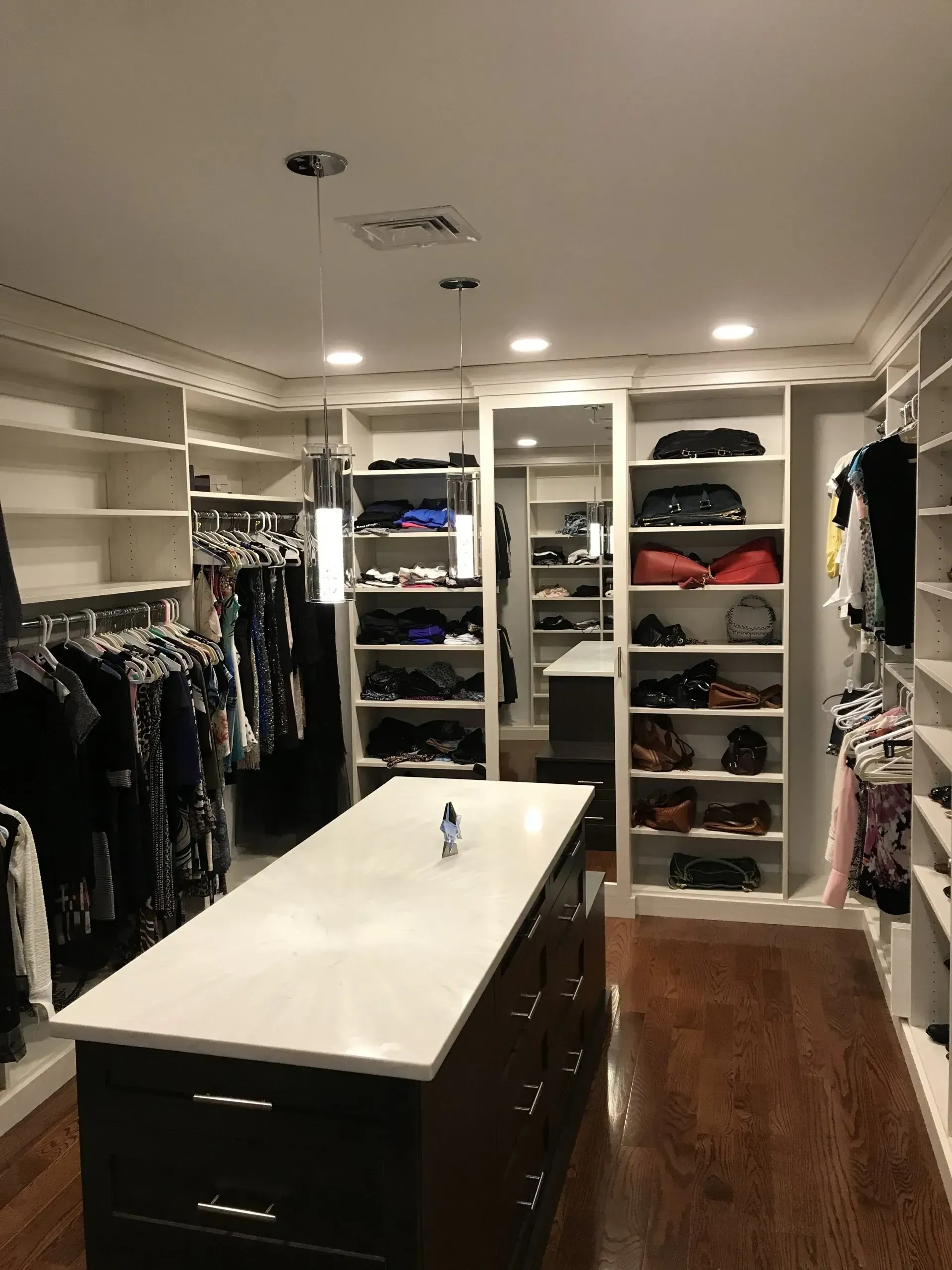 Walk-in closet with white shelves and a dark island. Clothes, shoes, and drawers. Wooden floor, neutral walls.