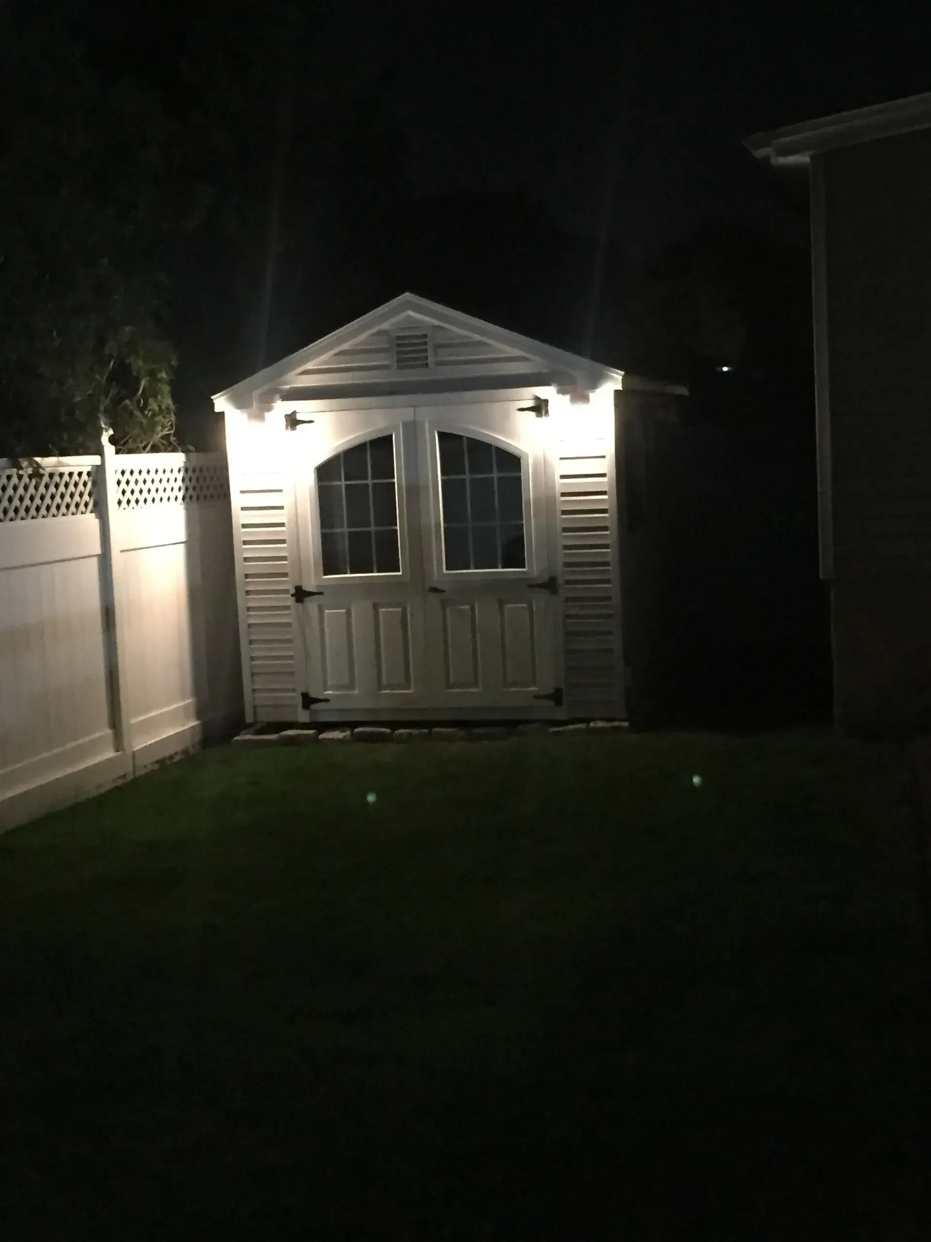 White shed with double doors and lights, in a yard at night, with white fence.