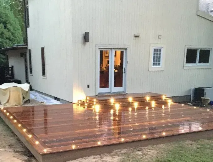 Wooden deck with string lights, attached to a light-colored house with glass doors.