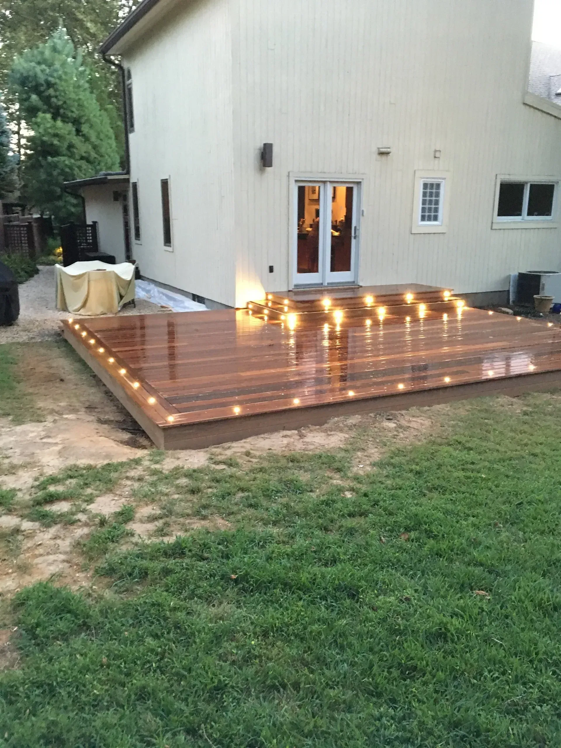 Wooden deck with built-in lights, attached to a light-colored house with glass doors.
