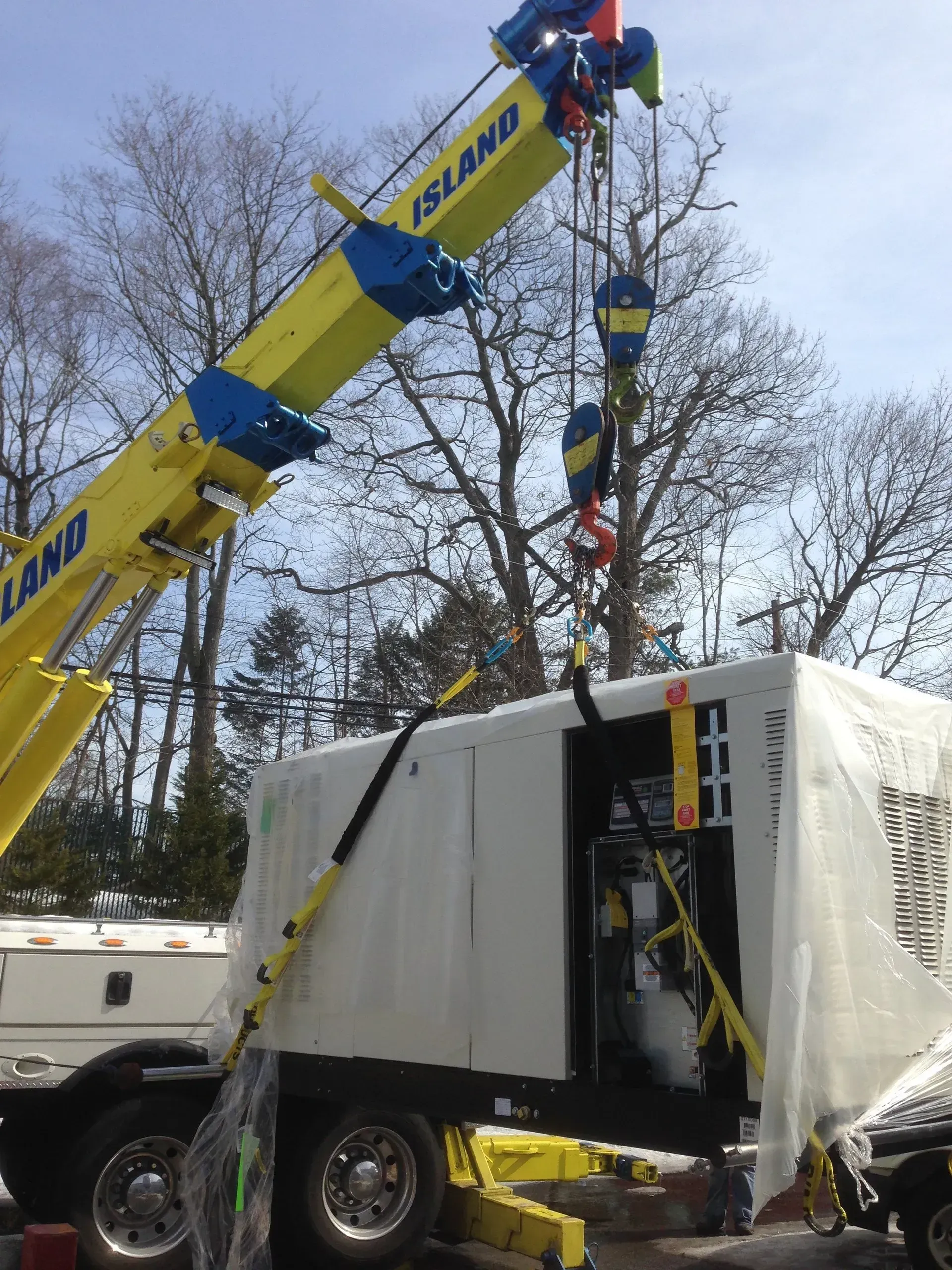 Yellow crane lifting a white generator on a trailer. Blue sky, trees in the background. 