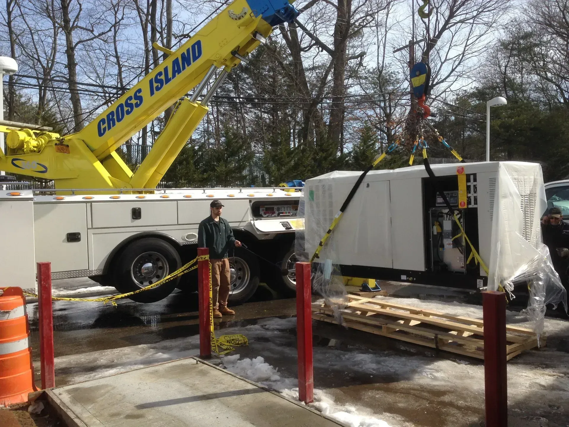 A yellow crane lifts a generator on a pallet. A man stands nearby.